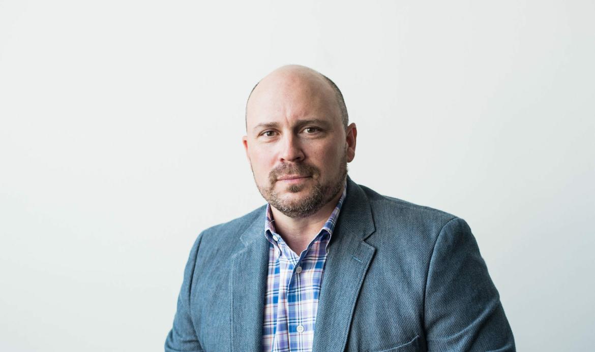 A man in a blazer and checkered shirt sitting against a white background.