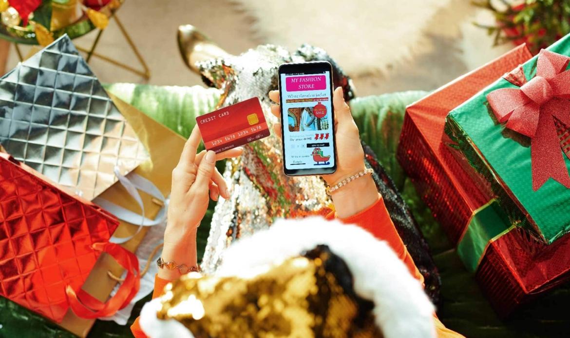 Person holding a credit card and a smartphone with a shopping app on screen, surrounded by colorful gift boxes.