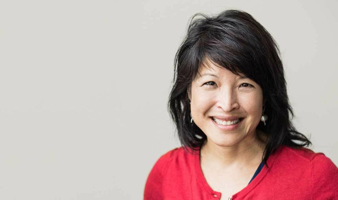 A smiling woman with black hair wearing a red top against a light grey background.