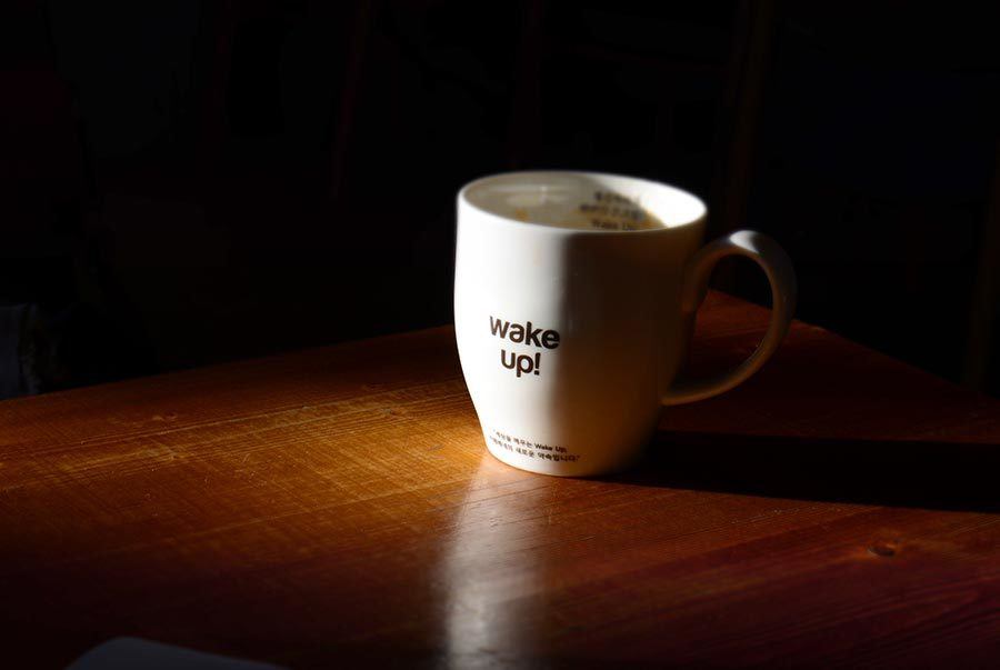 A white mug with the text "wake up!" on a wooden surface, partially illuminated by a beam of sunlight.