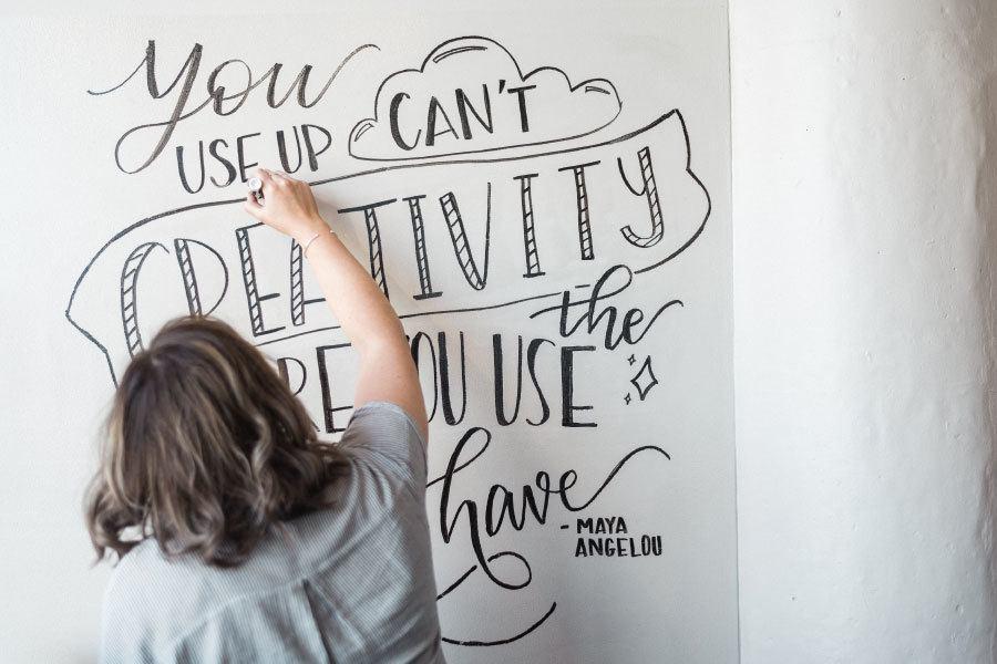 A person lettering a quote about creativity on a wall.