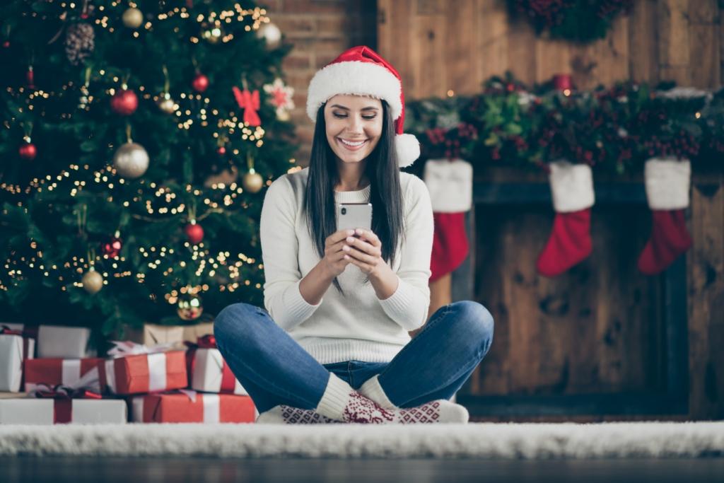 Full length photo of positive girl sir comfort floor carpet use her smartphone type text messages search christmas discounts wear santa claus hat in house with x-mas decoration indoors Full length photo of positive girl sir comfort floor carpet use her smartphone type text messages search christmas discounts wear santa claus hat in house with x-mas decoration indoors