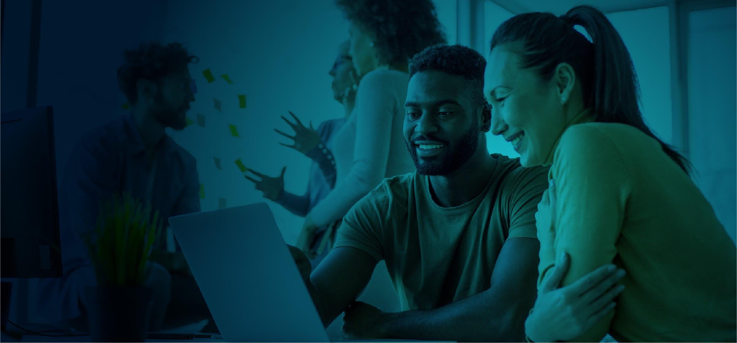 Two colleagues working on a laptop in a blue-toned office environment with others collaborating in the background.