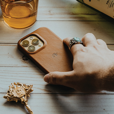 A person's hand rests on a wooden table next to a leather-covered smartphone with a Bullstrap case and a glass of amber liquid.