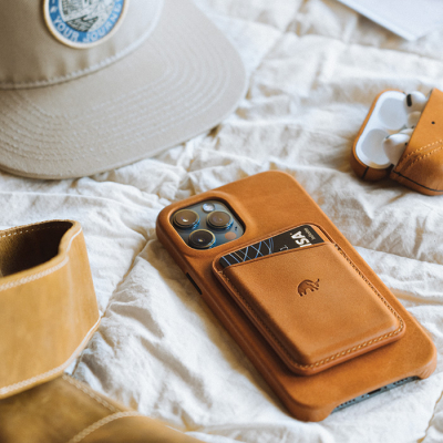 Leather Bullstrap smartphone case with earbuds on a fabric surface next to a cap and wallet.